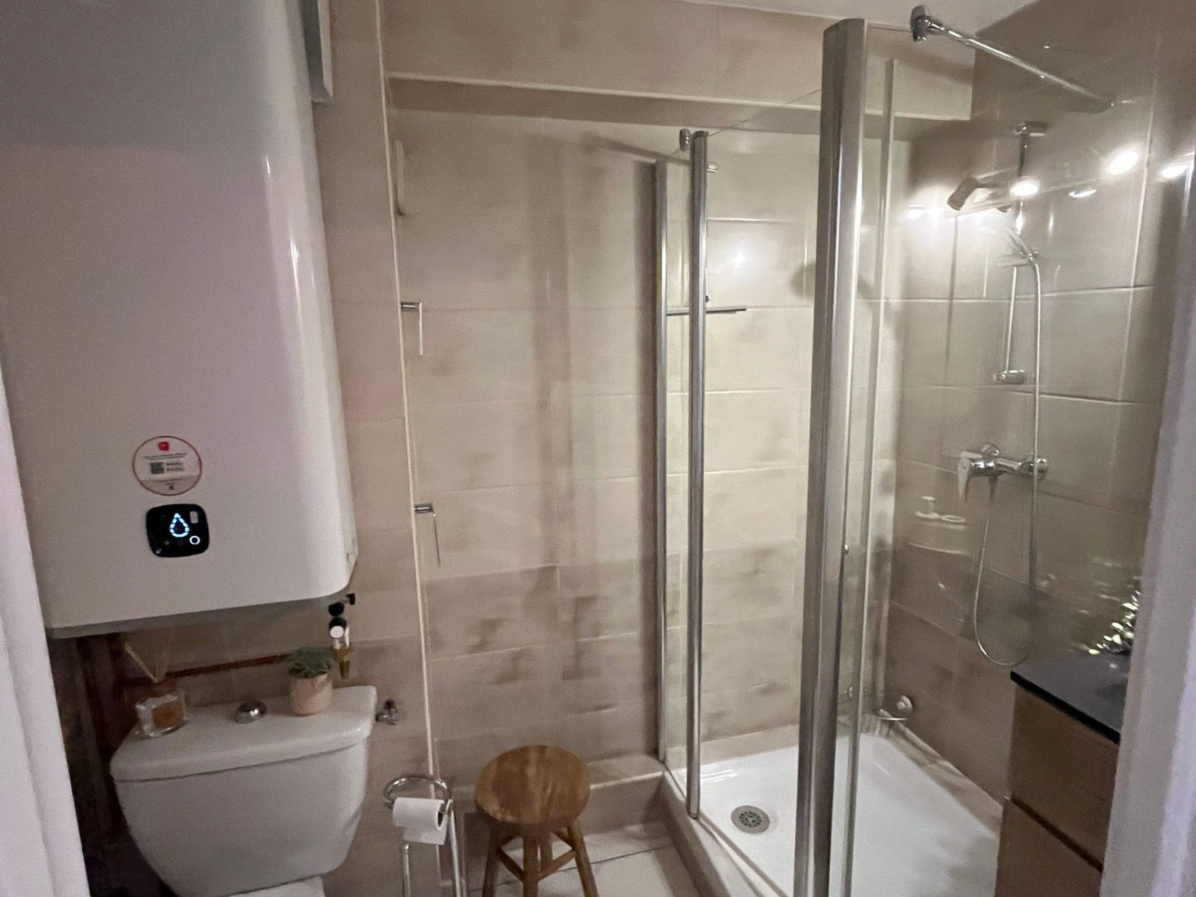 A modern bathroom with a glass-enclosed shower, a toilet, and a small wooden stool.