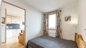 Ein Schlafzimmer mit einem hölzernen Bett, einem Fenster mit blumigen Vorhängen sowie Blick auf eine Küche und einen Essbereich.