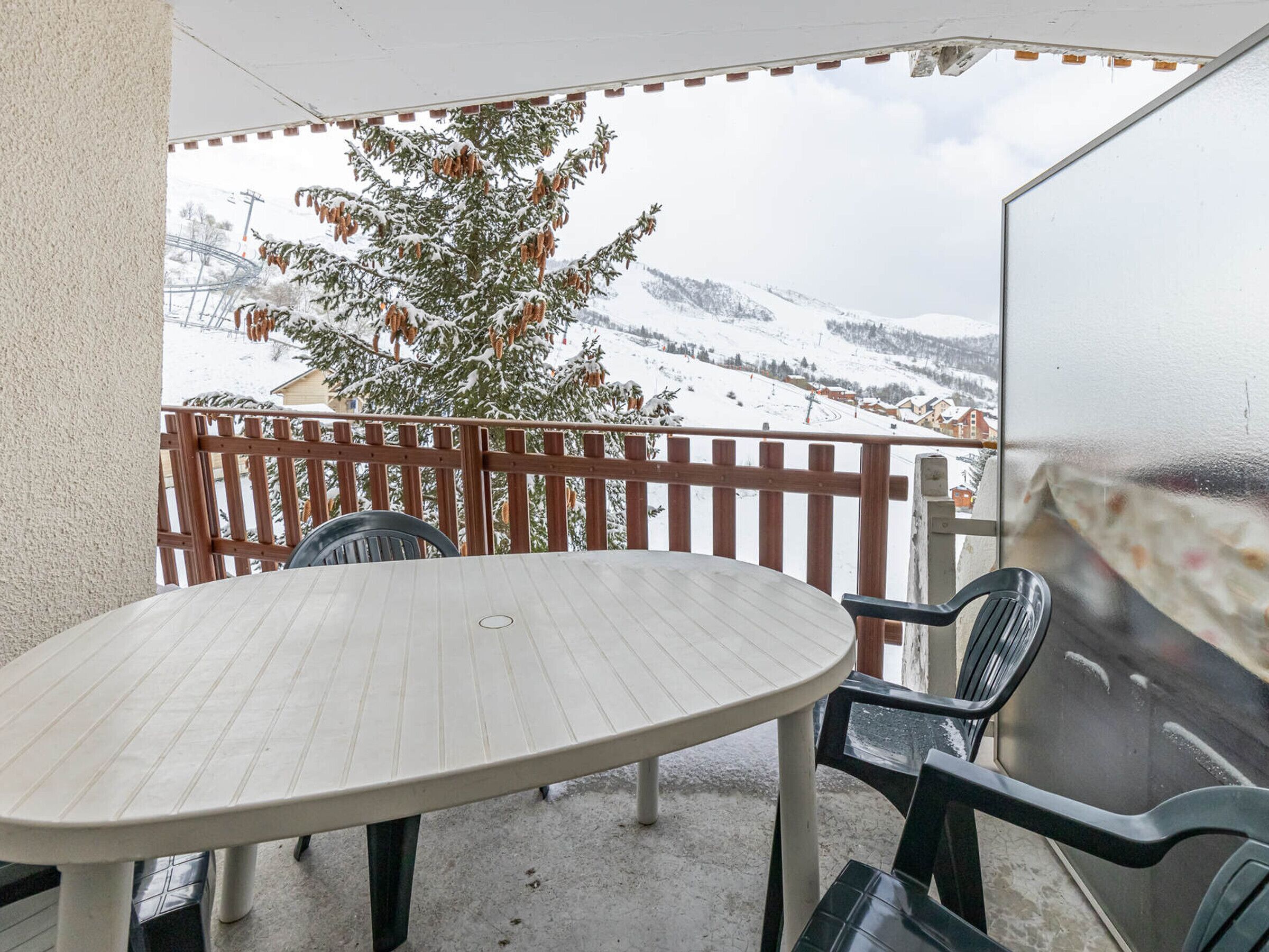 A balcony with a table and chairs, overlooking a snowy landscape with a tree and houses.