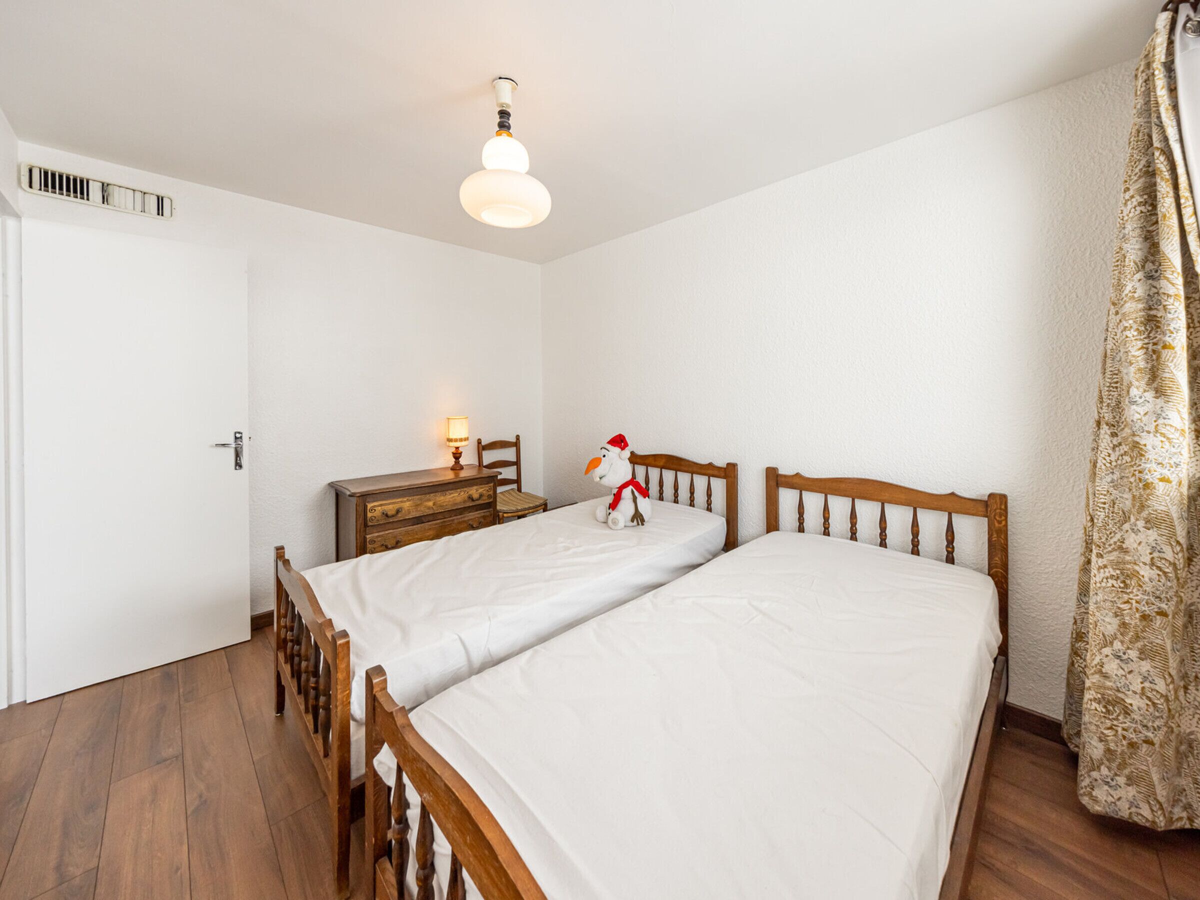 A bedroom with two wooden beds, a nightstand, a lamp, a chair, and a window with curtains.