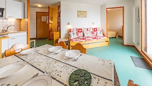 A compact room with a bed, a small table set for dining, a kitchenette, and a door leading to another room.
