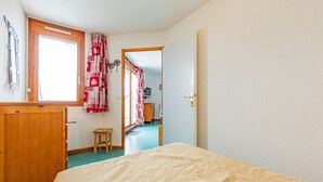A bedroom with a bed, a wooden dresser, a small stool, and a door leading to another room.