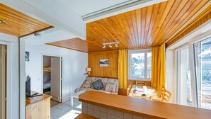 A compact kitchen with wooden cabinets and a central island, a dining area with a table and chairs, and a cozy seating area with a sofa and a window with curtains.