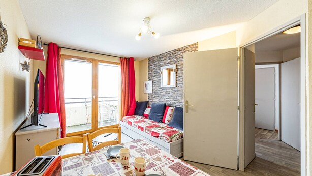 A cozy room with a dining table set for two, a small kitchenette, a bed with a quilt, and a balcony with red curtains. - Modern 2-bedroom apartment at the foot of the slopes (Saint-François-Longchamp)