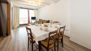 A dining area with a table set for breakfast, wooden chairs, a television in the background, and a balcony with a view.