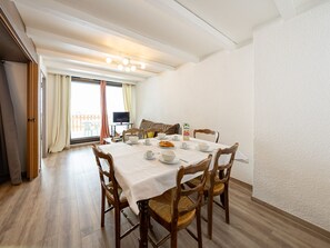 Uma área de refeições com mesa posta para o café da manhã, cadeiras de madeira, televisão ao fundo e uma varanda com vista.