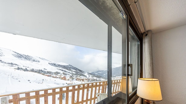 A room with a window overlooking a snowy mountain landscape, a wooden railing, and a table with a lamp.