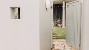 Un sendero estrecho que lleva a un patio trasero cercado, con un letrero con el número 22, un ventanal pequeño y una silla de mimbre.