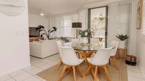 Una sala moderna con una mesa de vidrio, sillas blancas, un sofá y una cocina al fondo.