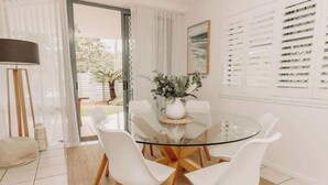 Un comedor moderno con una mesa de vidrio rodeada de sillas blancas, un florero con follaje y vistas al exterior a través de grandes ventanas con persianas.