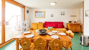 A dining area with a wooden table, chairs, and a bed with red pillows. There is a window with a view, a clock on the wall, and a microwave on a wooden cabinet. - Comfortable apartment 7 ppl near slopes and shops (Saint-François-Longchamp)