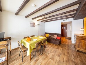 Uma área de refeições com uma mesa posta para quatro pessoas, piso de madeira, um sofá marrom com almofadas e uma televisão sobre um armário de madeira.