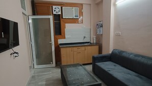 Ein kompakter Wohnraum mit Deckenventilator, einer Küchenzeile mit Mikrowelle, einem Glastisch, einer Couch und einem an der Wand befestigten Fernseher.
