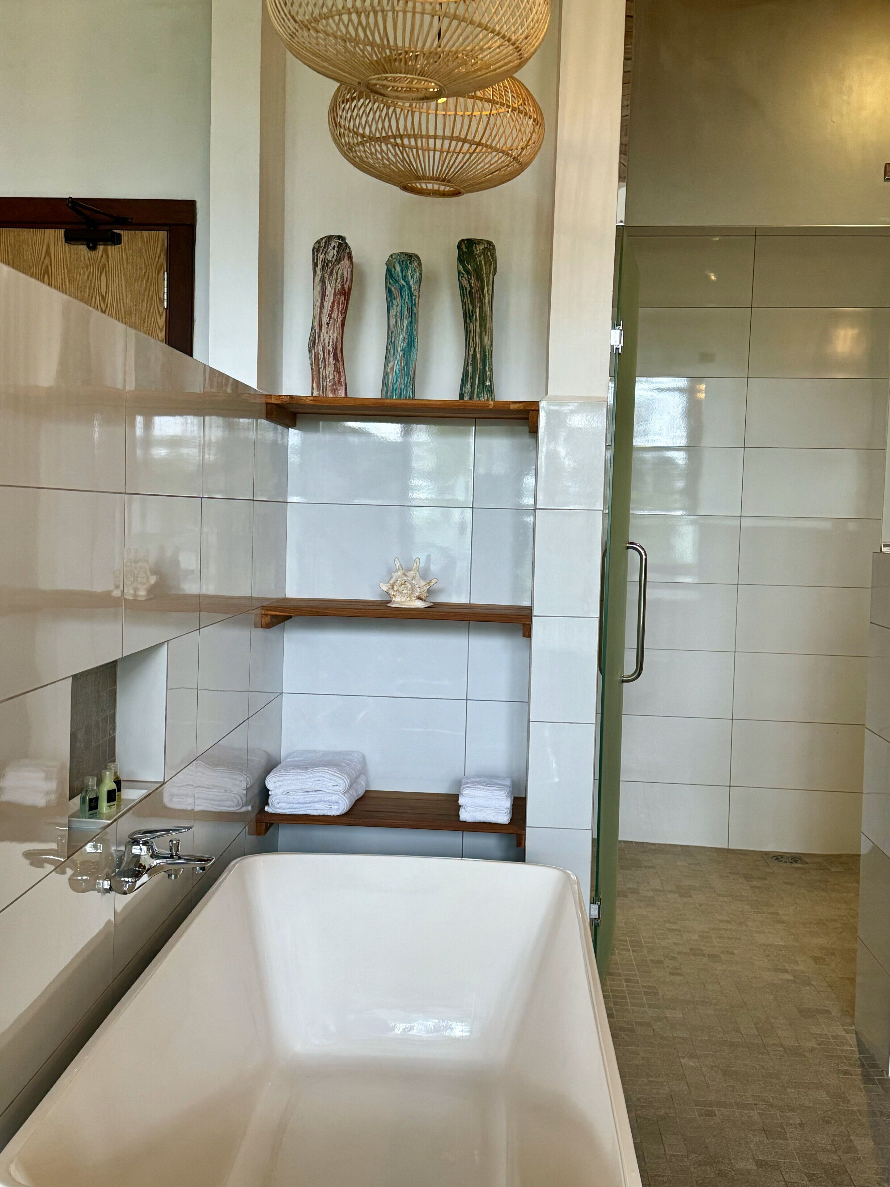 A modern bathroom with a large bathtub, white tiled walls, and a wooden shelf with decorative items.