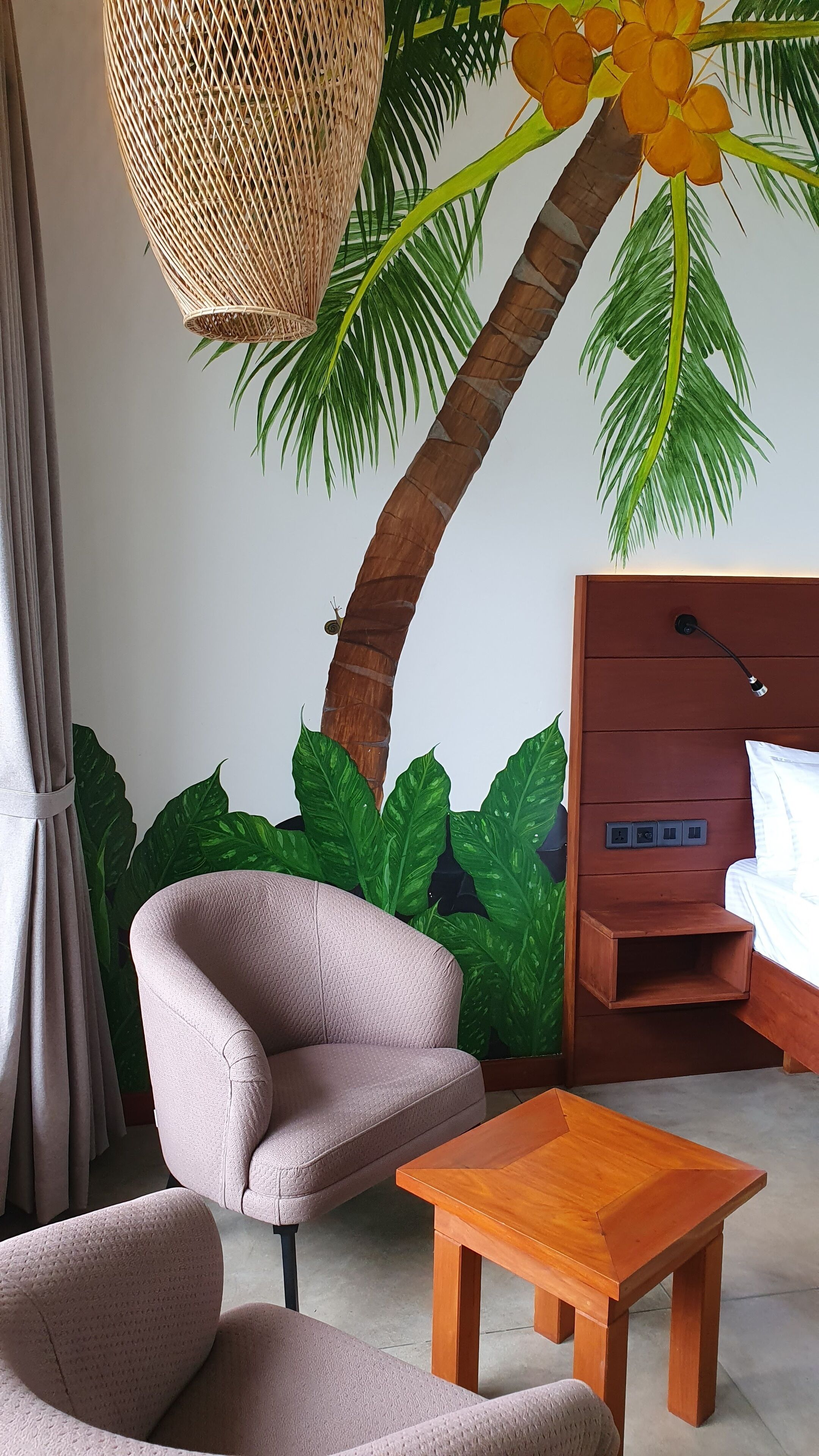 A room with a bed, a wooden nightstand, a lamp, a woven hanging light fixture, and a large artificial palm tree with green leaves and orange flowers.