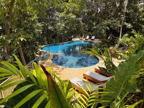 Una piscina circular rodeada de abundante vegetación y tumbonas de madera.