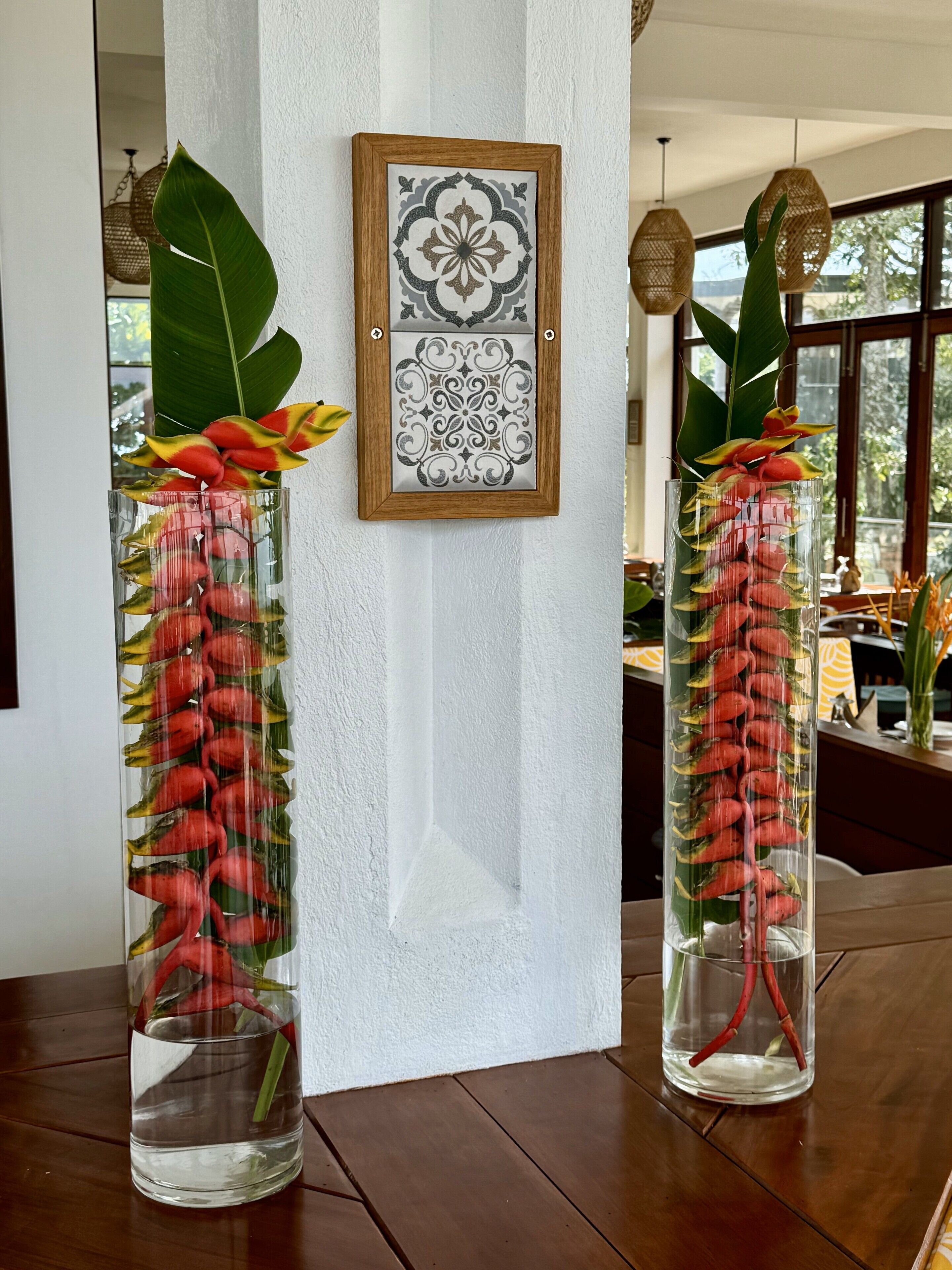 Two tall glass vases with red flowers and green leaves on a wooden table, with a decorative wall panel and hanging lights in the background.
