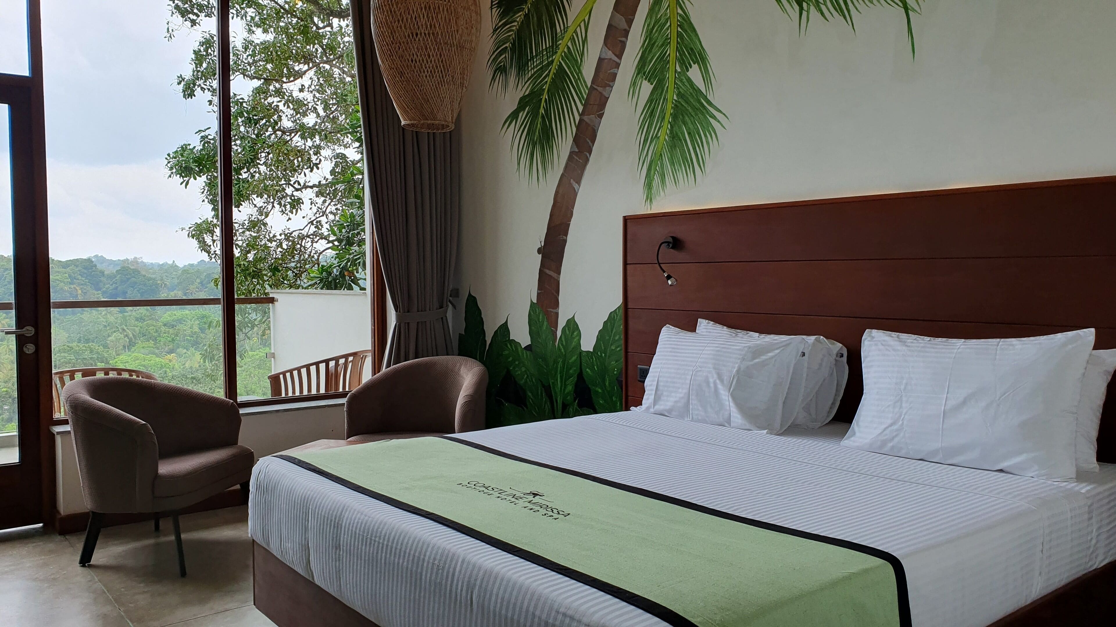 A hotel room with a large bed, a chair, a view of the outdoors, and a tropical-themed wall.