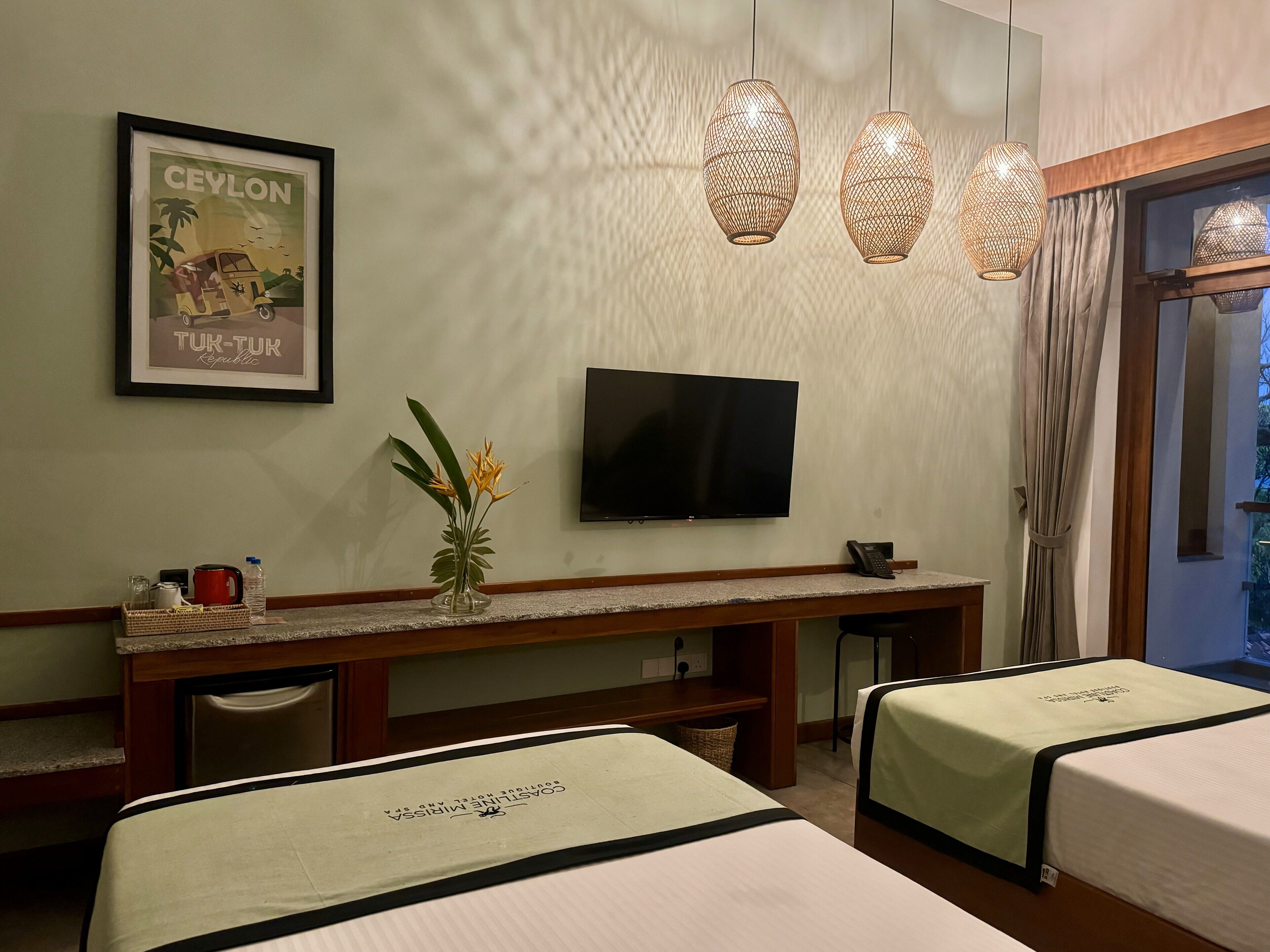 A hotel room with two beds, a TV, a desk with a vase of flowers, and a framed picture on the wall.