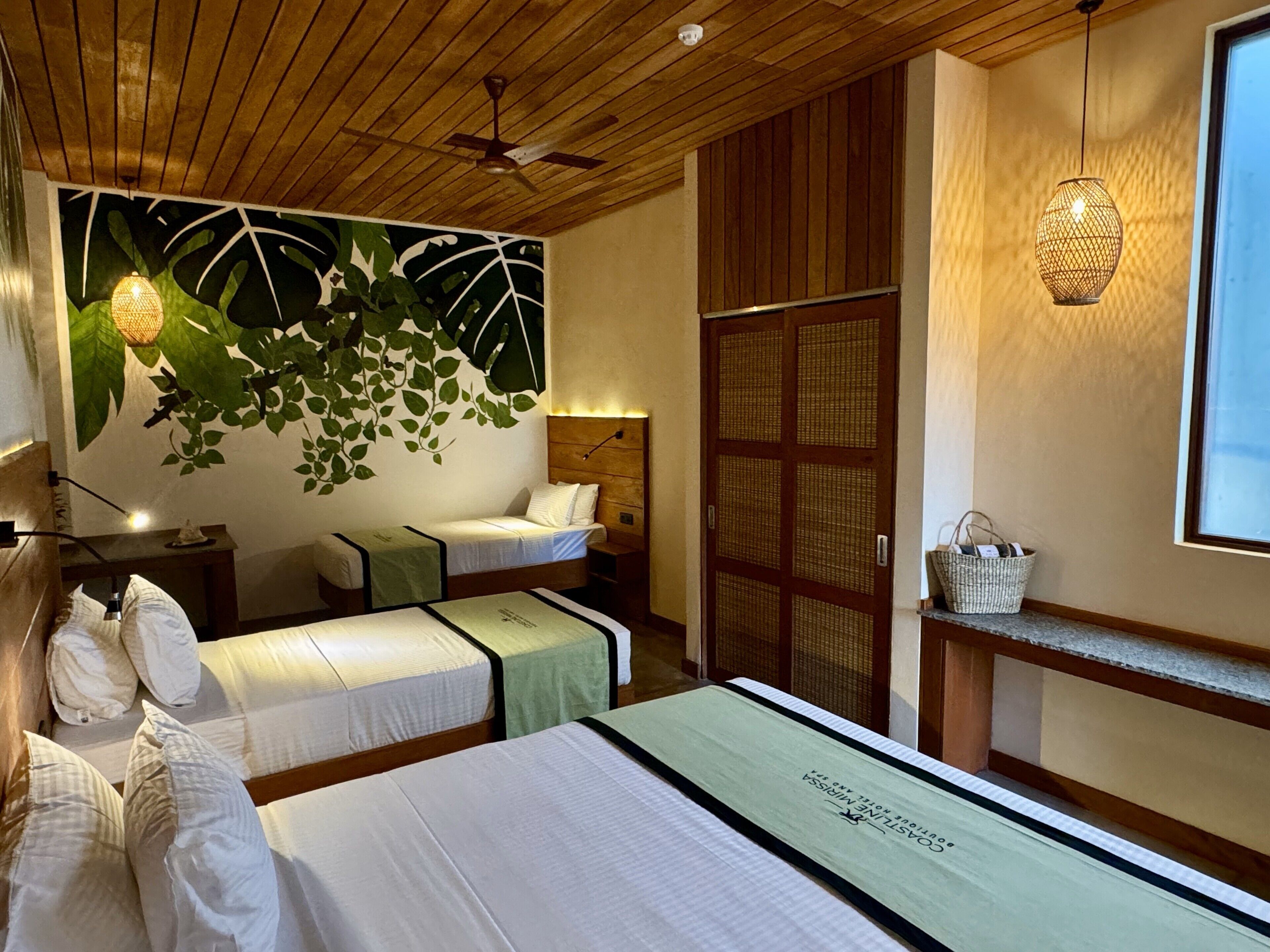 A hotel room with two beds, a wooden ceiling, a wall mural, a traditional woven lamp, and a window with a view.