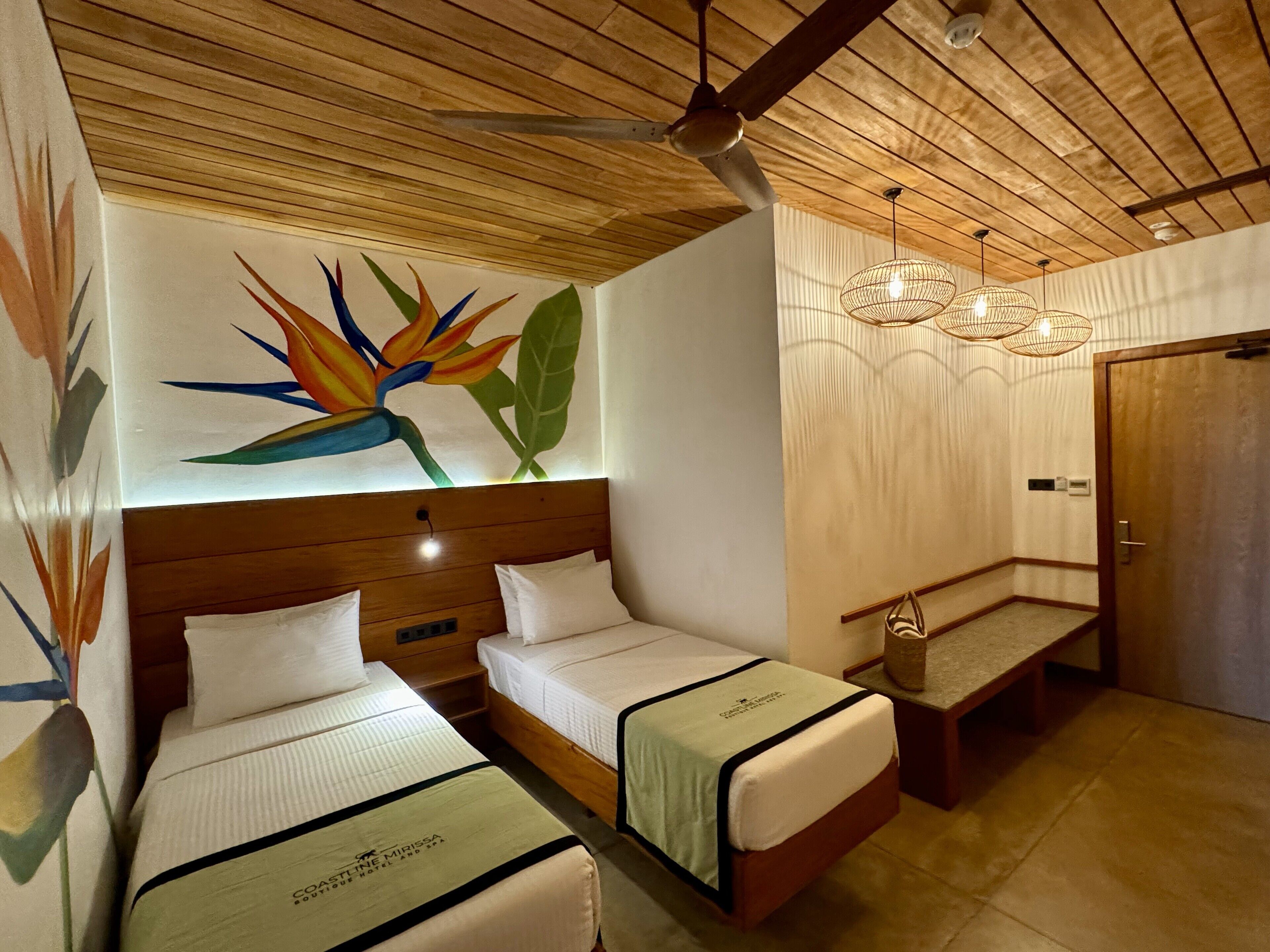 A hotel room with two beds, a wooden ceiling, a colorful wall mural, and a small seating area.