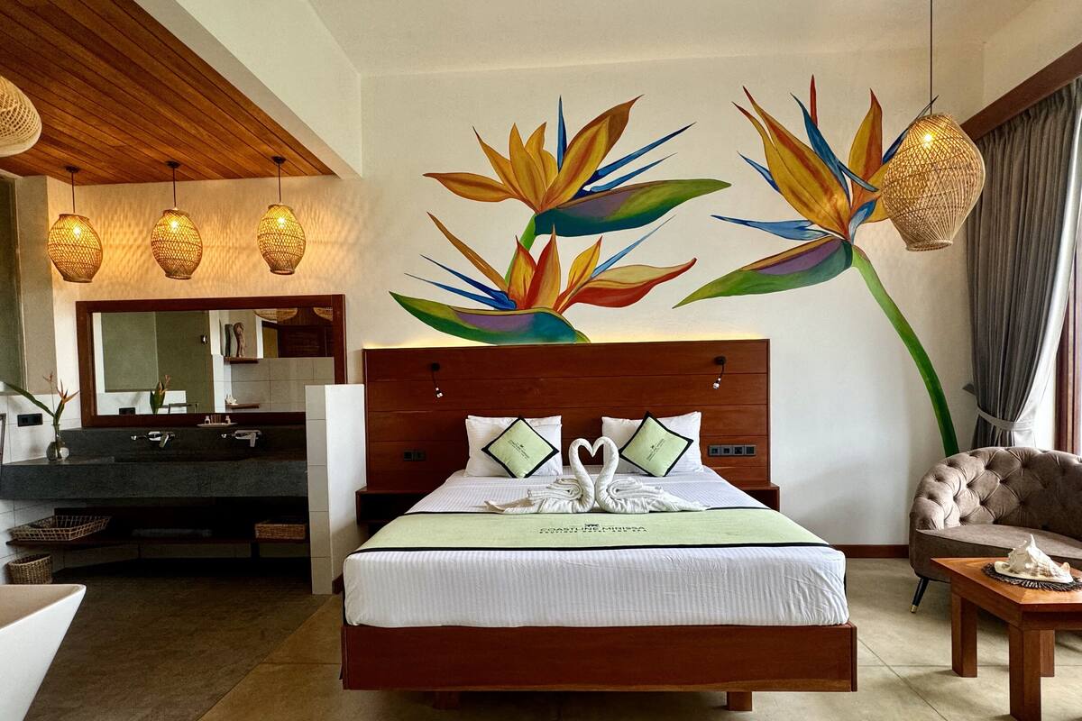 Habitación de hotel con una cama grande, cabecero de madera, baño con espejo y lavamanos, y zona de comedor con mesa y sillas.