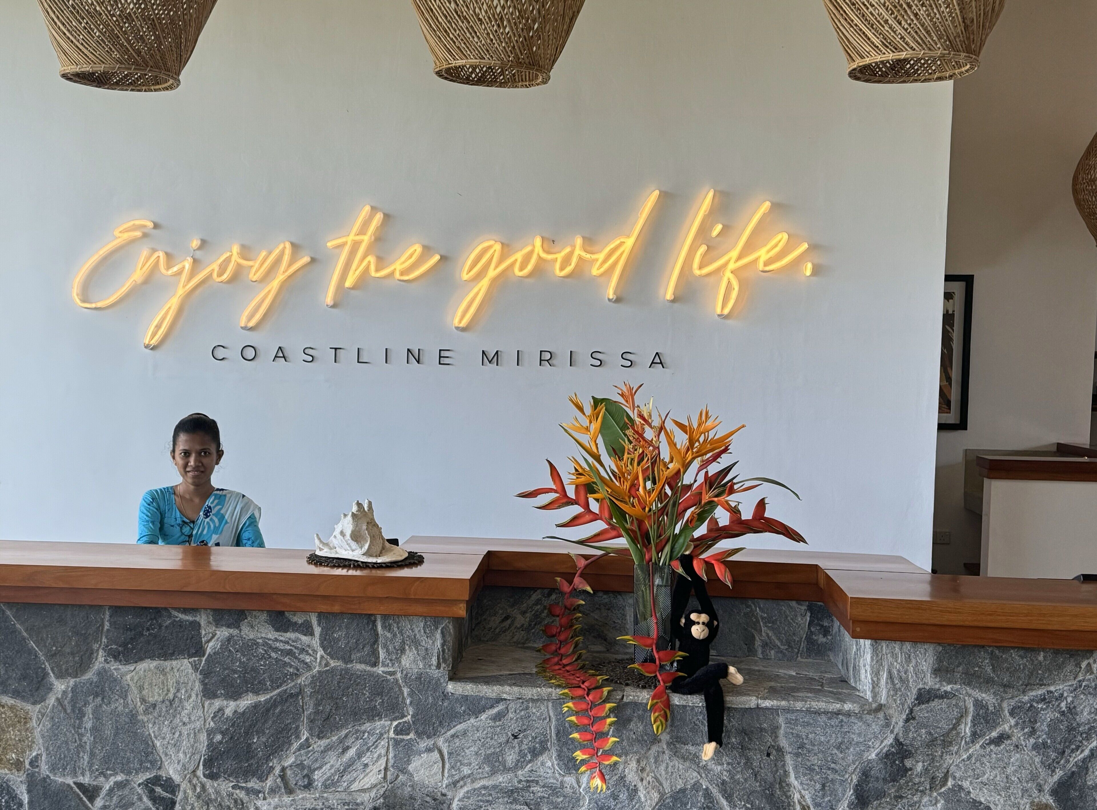 A reception desk with a neon sign that reads "Enjoy the good life." and "Coastline Mirissa". There is a woman seated behind the desk, a decorative plant, and a monkey figurine on the counter.