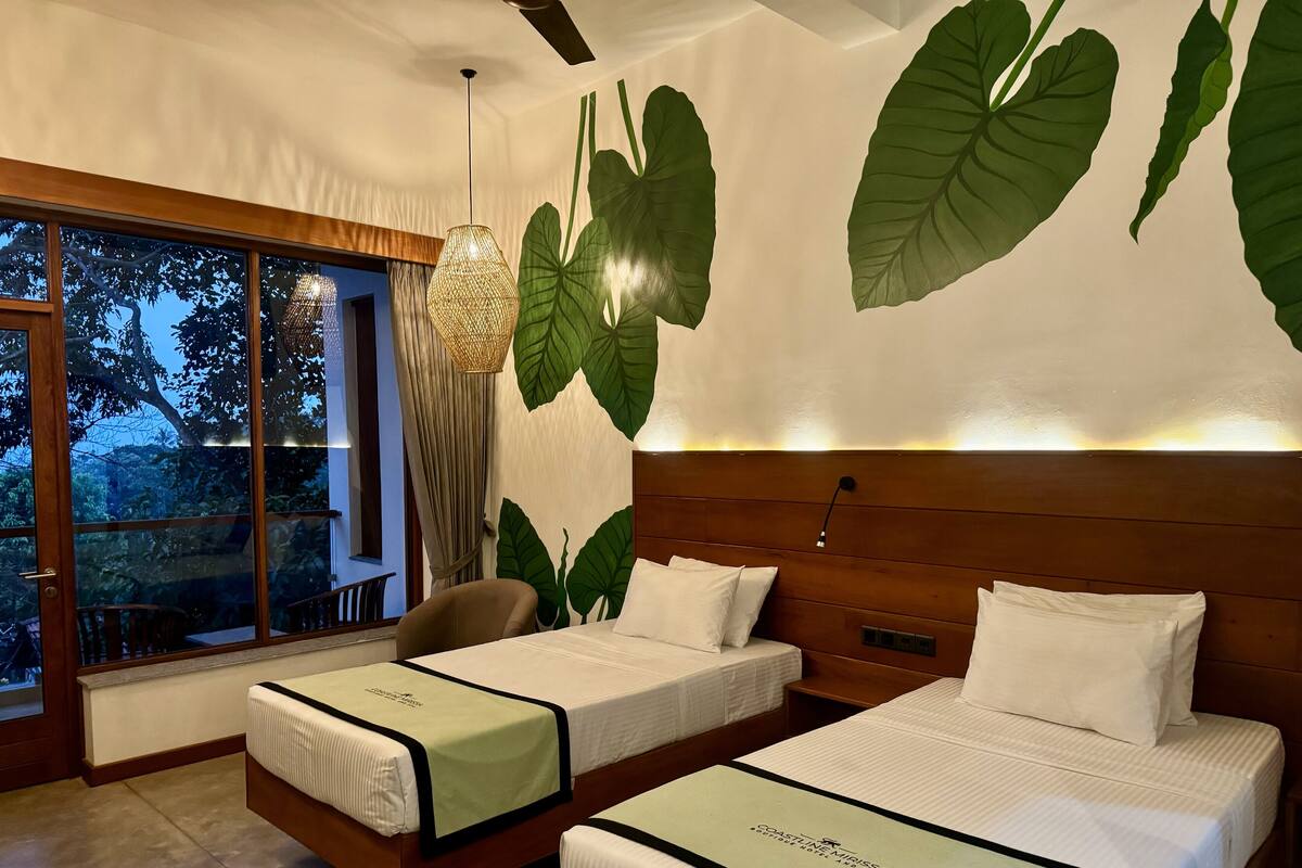Habitación de hotel con dos camas, un ventanal con vistas a árboles y una pared con hojas verdes grandes.