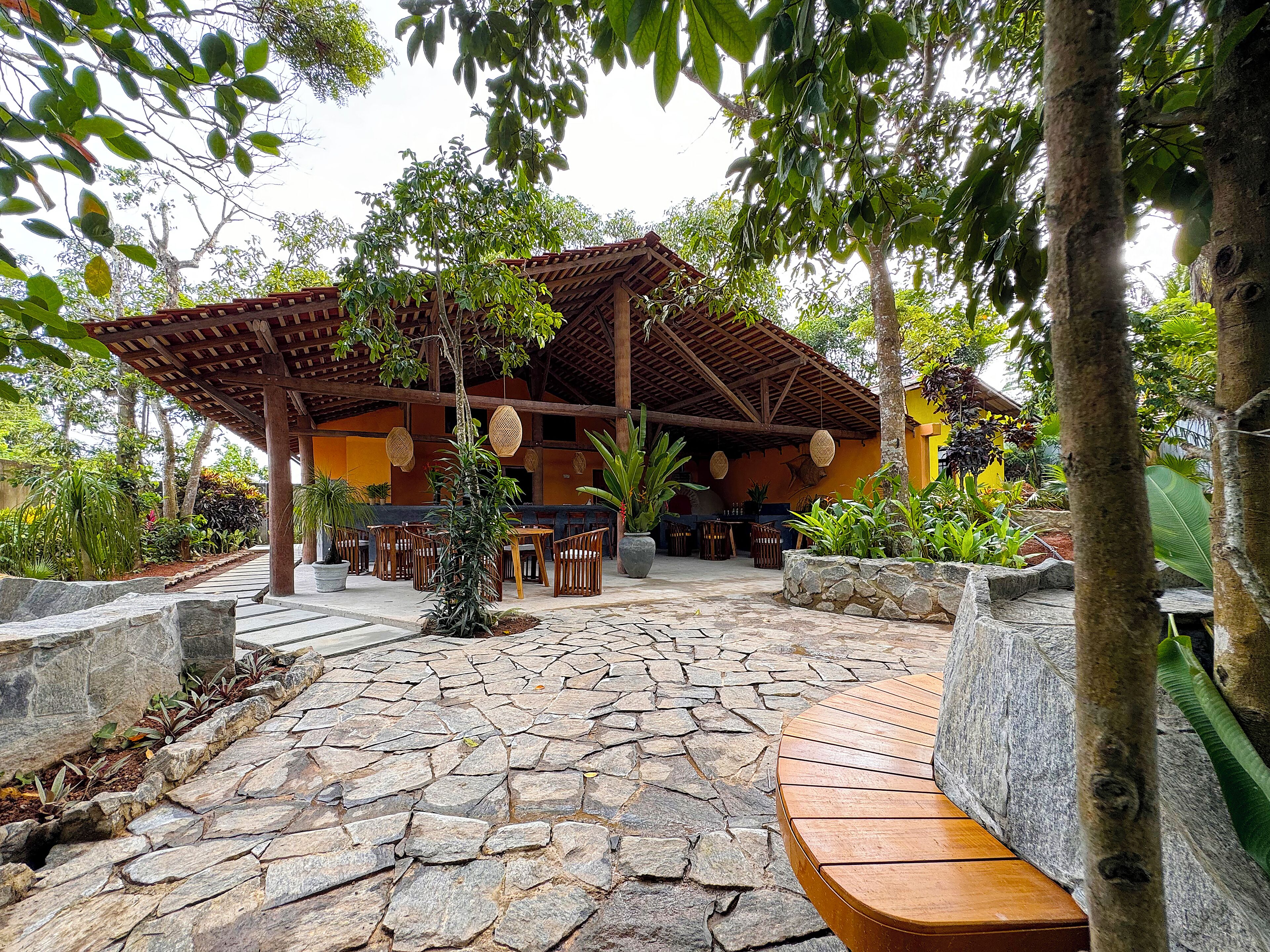 A covered outdoor seating area with stone flooring, wooden furniture, and lush greenery.