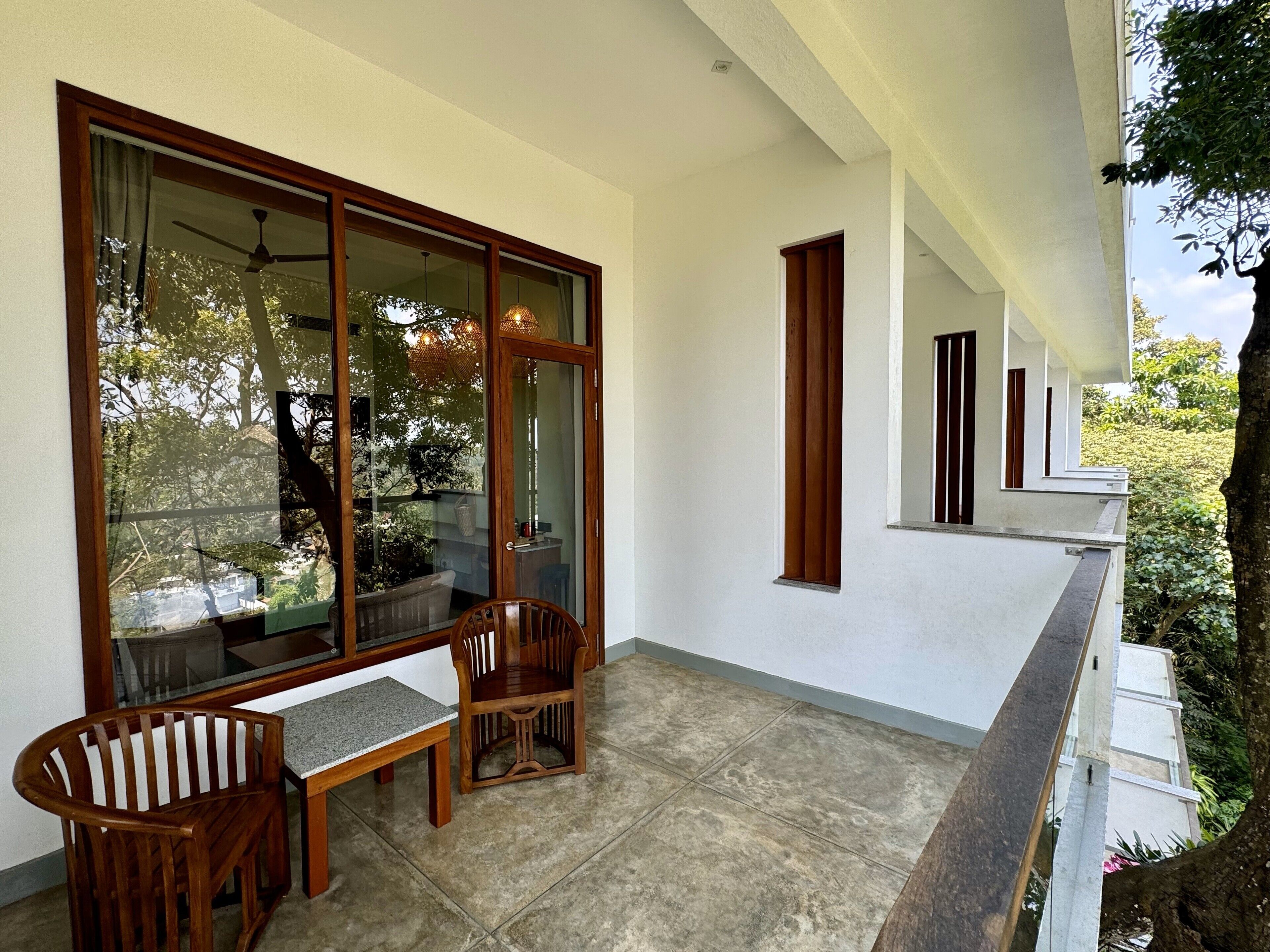 Un balcón con mesa y sillas de madera, ventanas amplias y vistas a árboles y un edificio.
