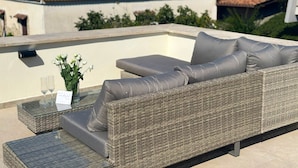 A wicker sofa with cushions on an outdoor terrace with a glass table and a vase of flowers. - Luxury Apartments Collection - The Sand Suite (Passoscuro)