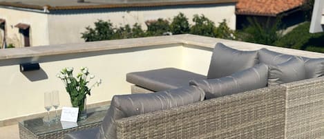 A wicker sofa with cushions on an outdoor terrace with a glass table and a vase of flowers.