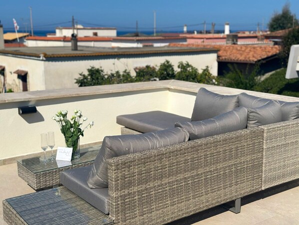 A wicker sofa with cushions on an outdoor terrace with a glass table and a vase of flowers.