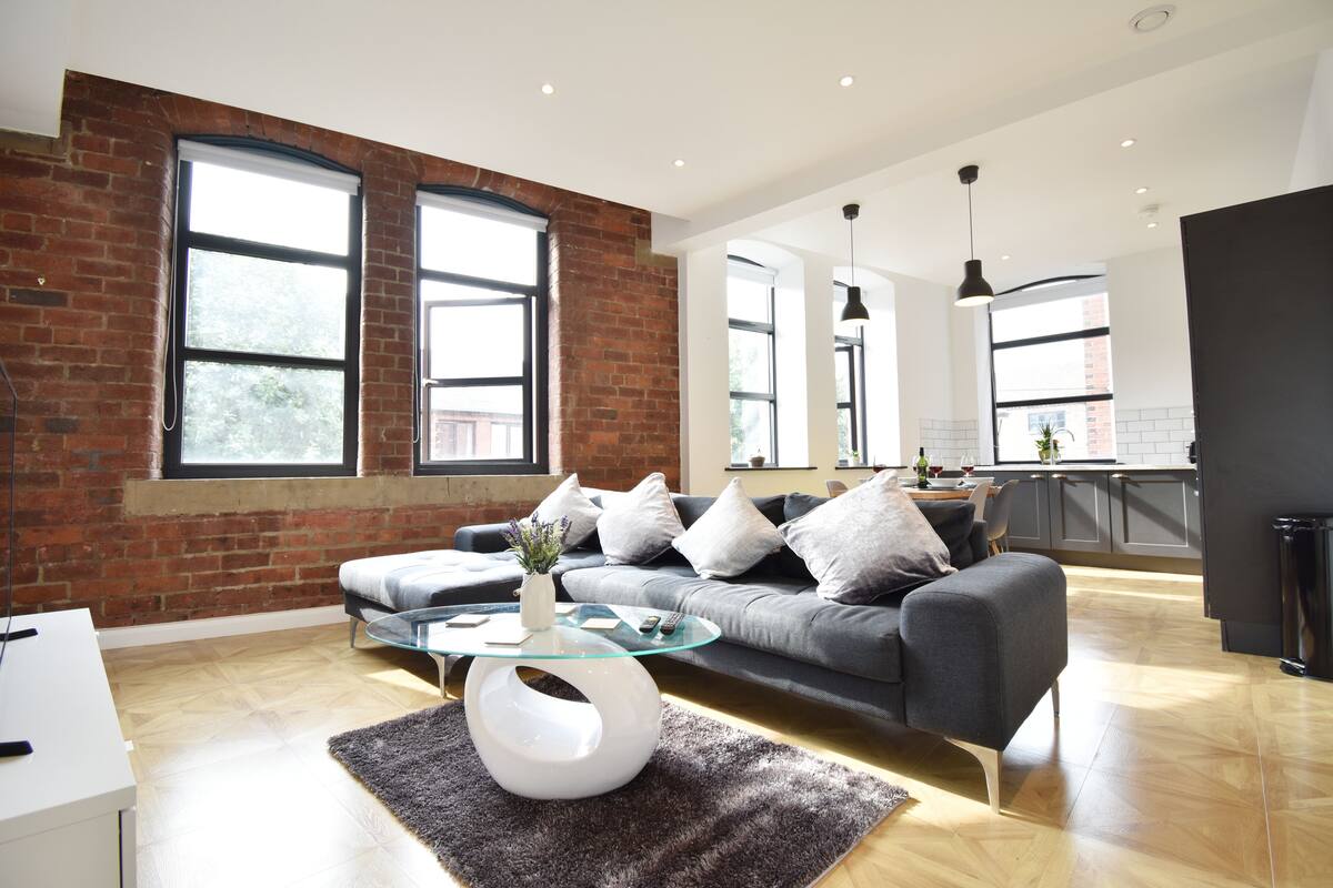 A modern living room with a grey sofa, a glass coffee table, and a brick wall.