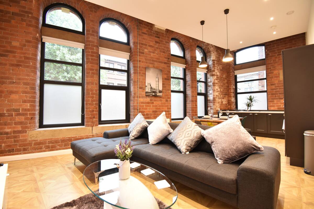 A modern living room with a grey sofa, a glass coffee table, and a brick wall with large windows.