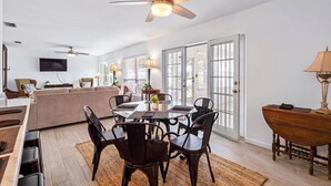A spacious living room with a glass dining table, dark wooden chairs, a ceiling fan, and a large window with blinds. - Bayfront Cottage -- Close to everything (St. Augustine)