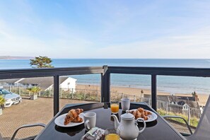 Una mesa de desayuno con cruasanes, una jarra de jugo de naranja y una taza, con vista a una playa con valla y un coche estacionado.