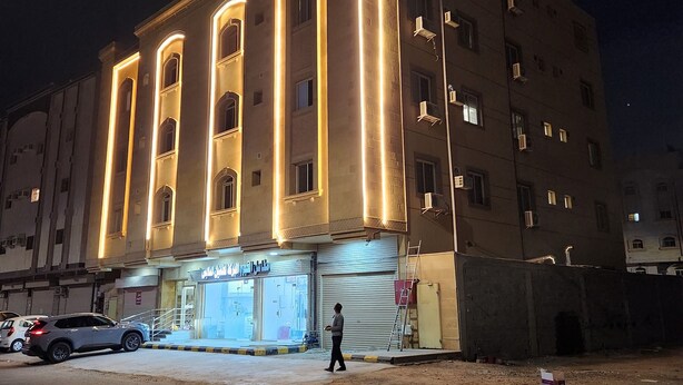 Un edificio de varios pisos con líneas verticales iluminadas en la fachada, un escaparate con señalización y una persona caminando por la acera.