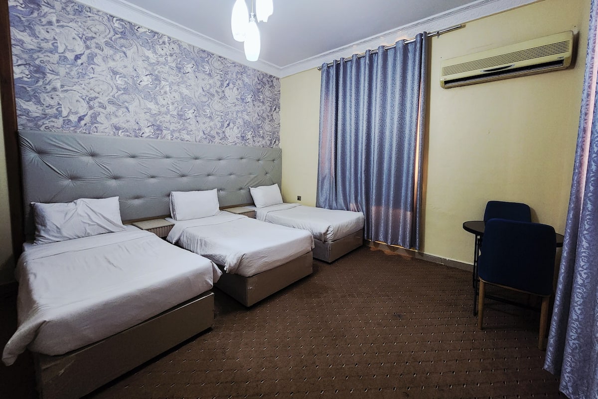 A hotel room with two single beds, a small desk, a chair, a patterned wall, and a ceiling light fixture.