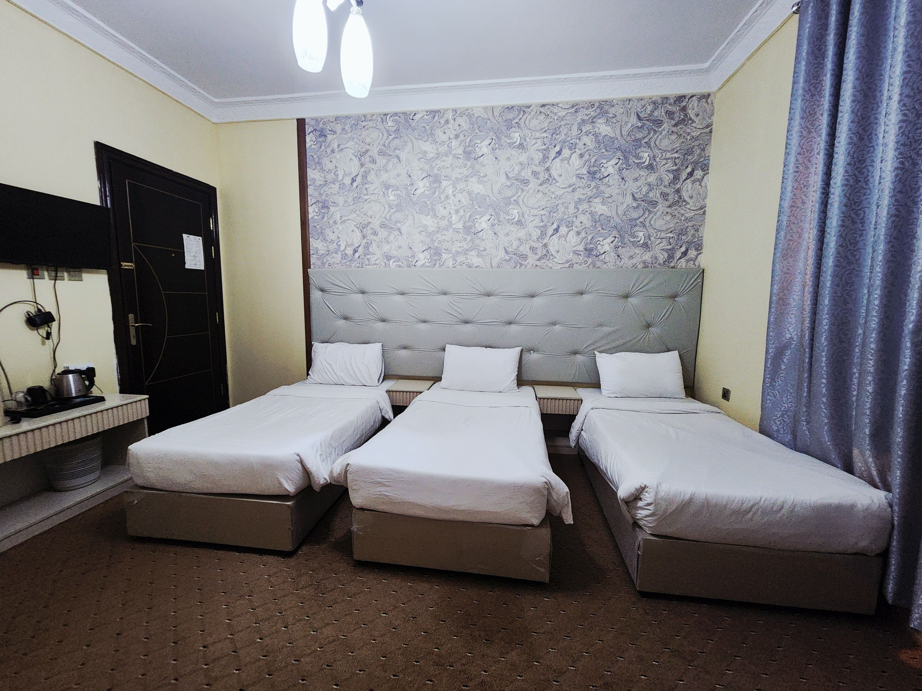 Habitación de hotel con tres camas individuales, un cabecero grande, una mesita pequeña, un espejo y una ventana con cortinas.
