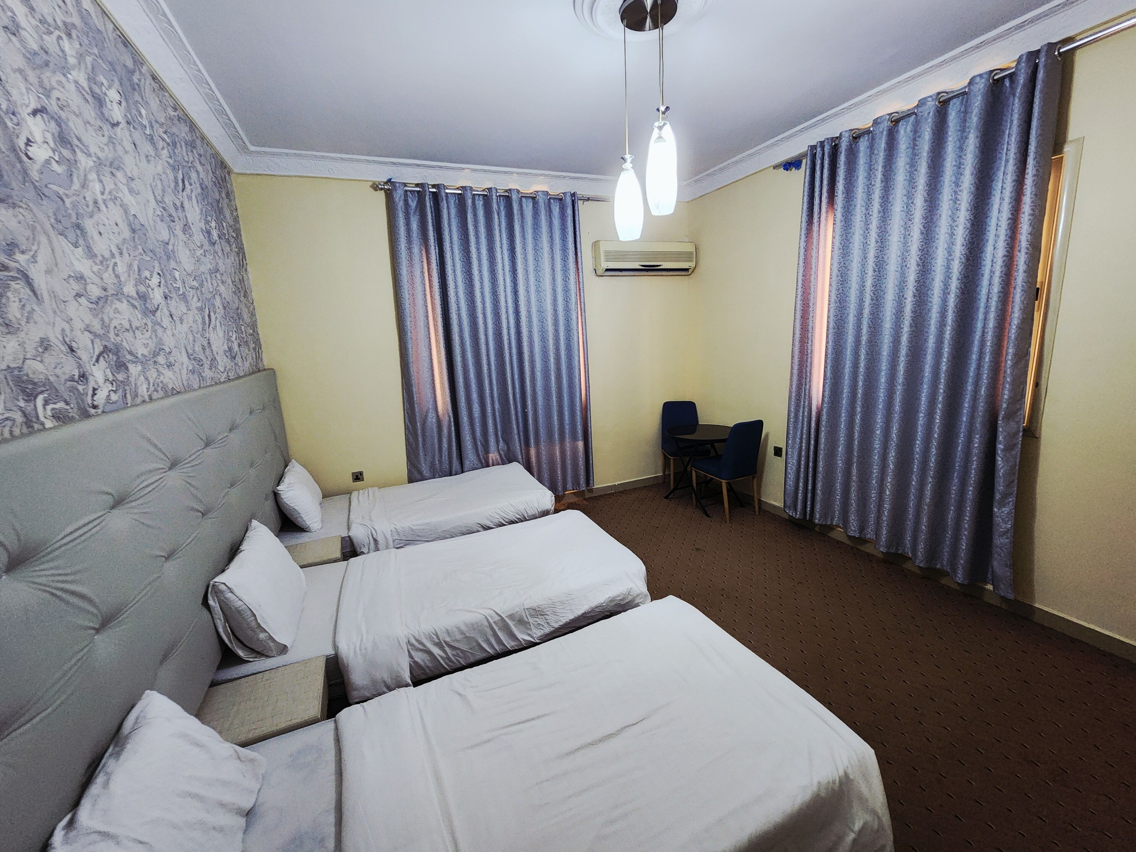 A hotel room with two single beds, a small table with two chairs, an air conditioning unit, and patterned curtains.