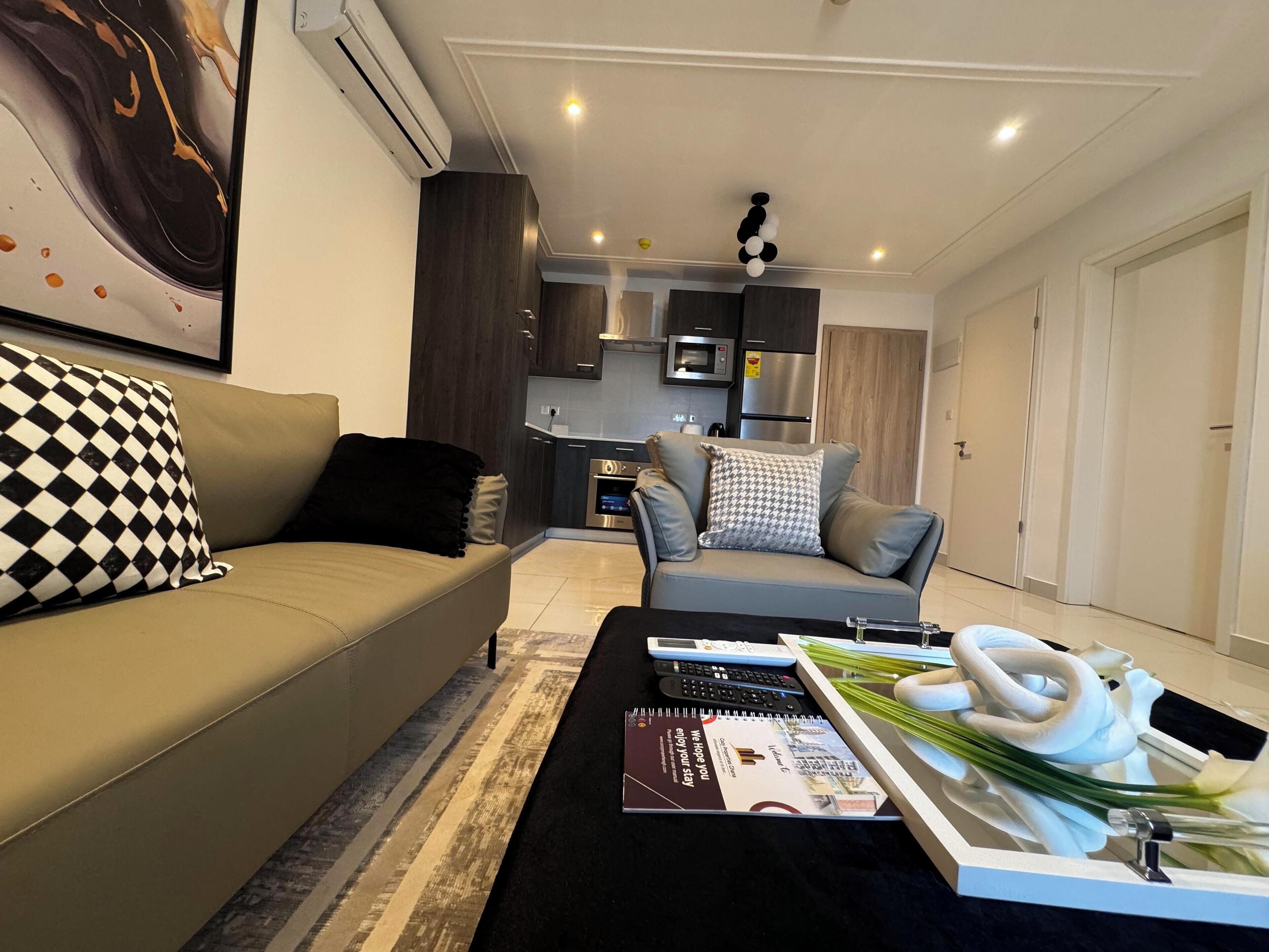 A modern living room with a sofa, a coffee table with remote controls and a magazine, and a kitchen area in the background.