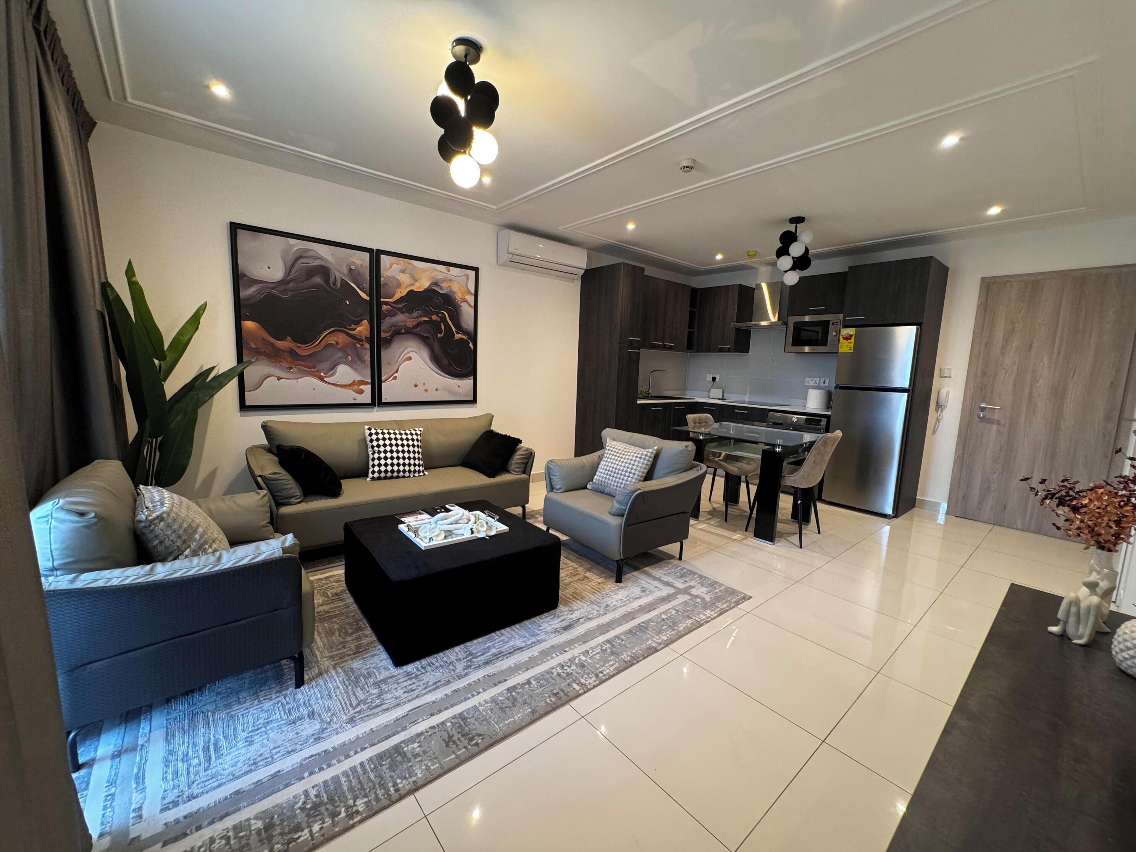A modern living room with a sofa, armchairs, a coffee table, and a dining area with chairs. There is a kitchen with dark cabinets and a refrigerator.