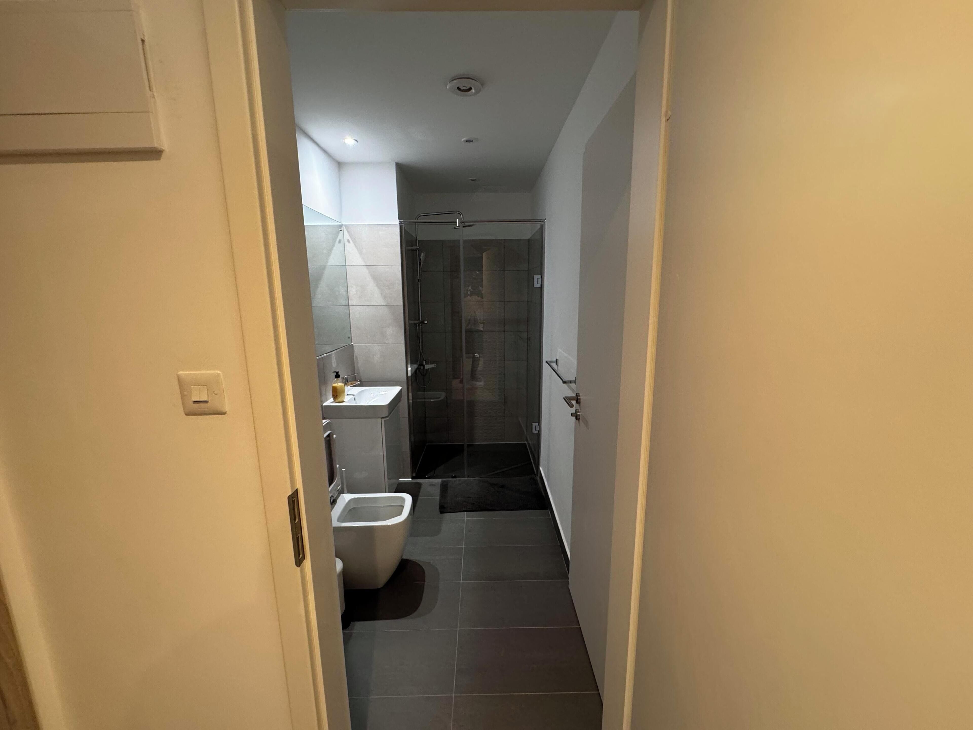 A narrow hallway leading to a bathroom with a toilet, bidet, and a glass-enclosed shower area.