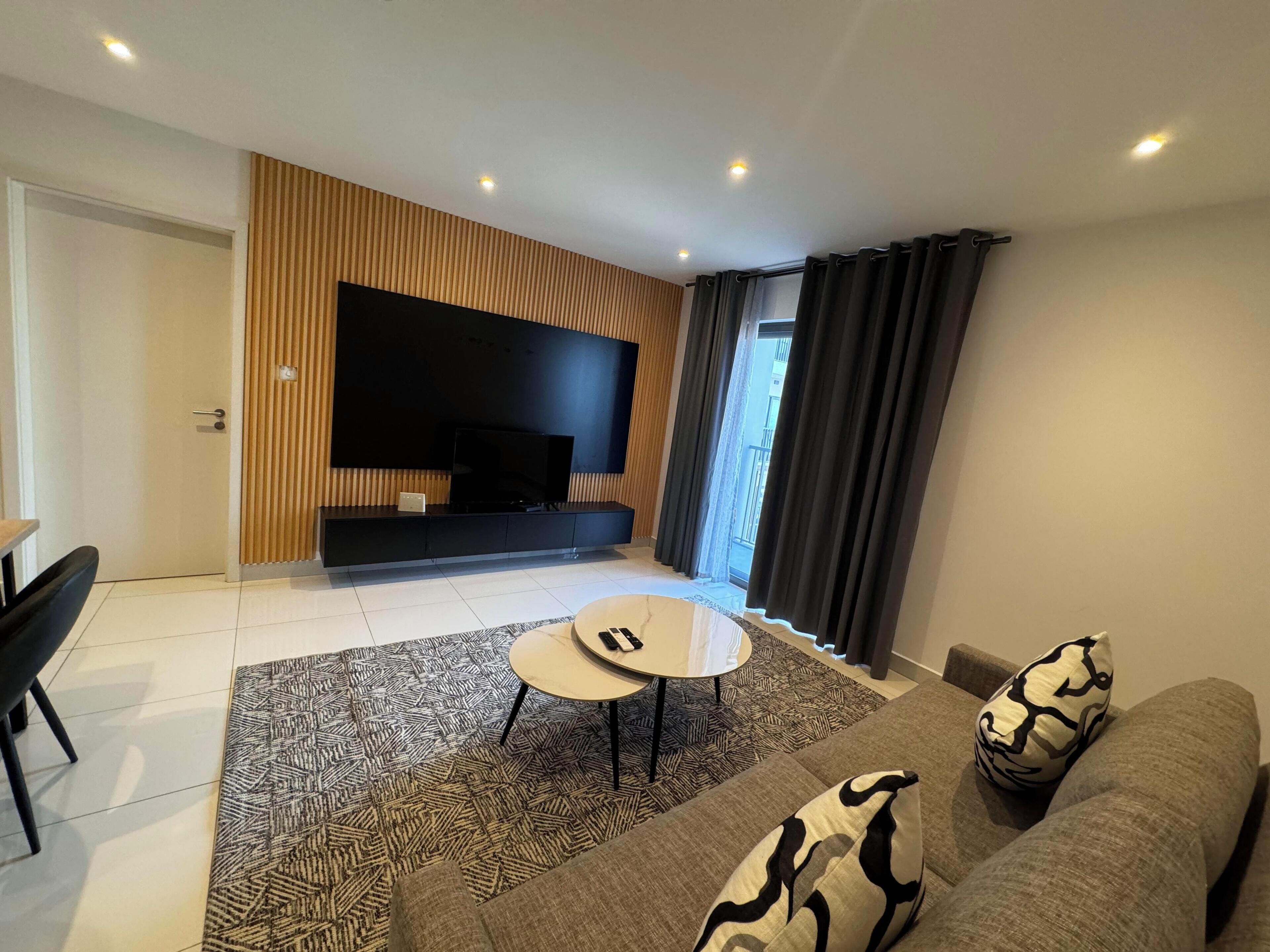 Een moderne woonkamer met een grote flatscreen-tv, een ronde salontafel en een bank met gekleurde kussens.