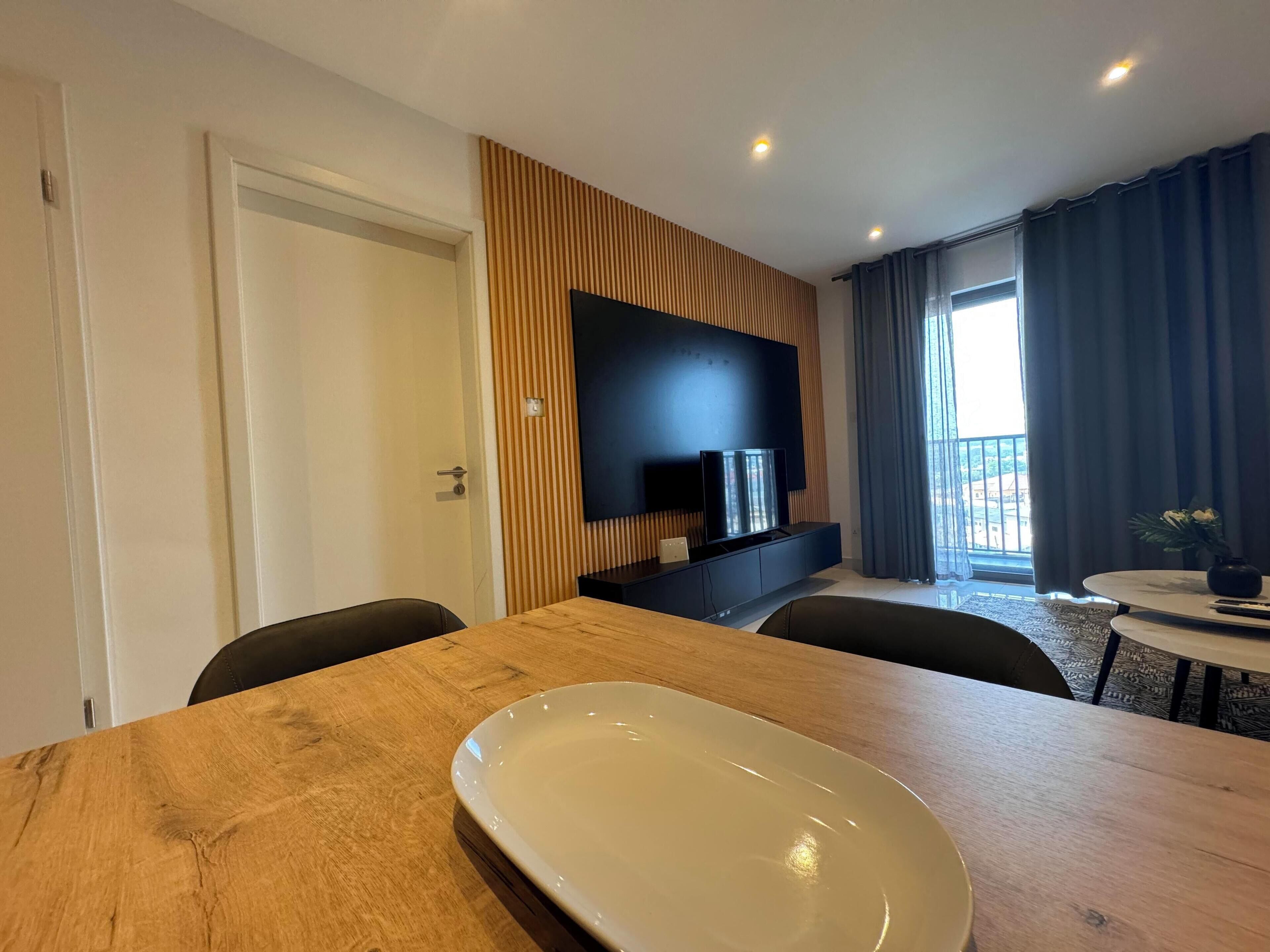 A modern hotel room with a wooden dining table, a flat-screen TV mounted on the wall, and a balcony with curtains.