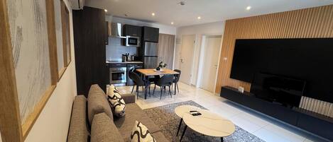 A modern living room with a sofa, coffee table, and TV. A kitchen with appliances and a dining area with chairs is visible in the background.