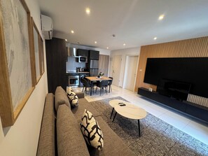 Ein modernes Wohnzimmer mit Sofa, Couchtisch und Fernseher. Im Hintergrund ist eine Küche mit Geräten und ein Essbereich mit Stühlen zu sehen.
