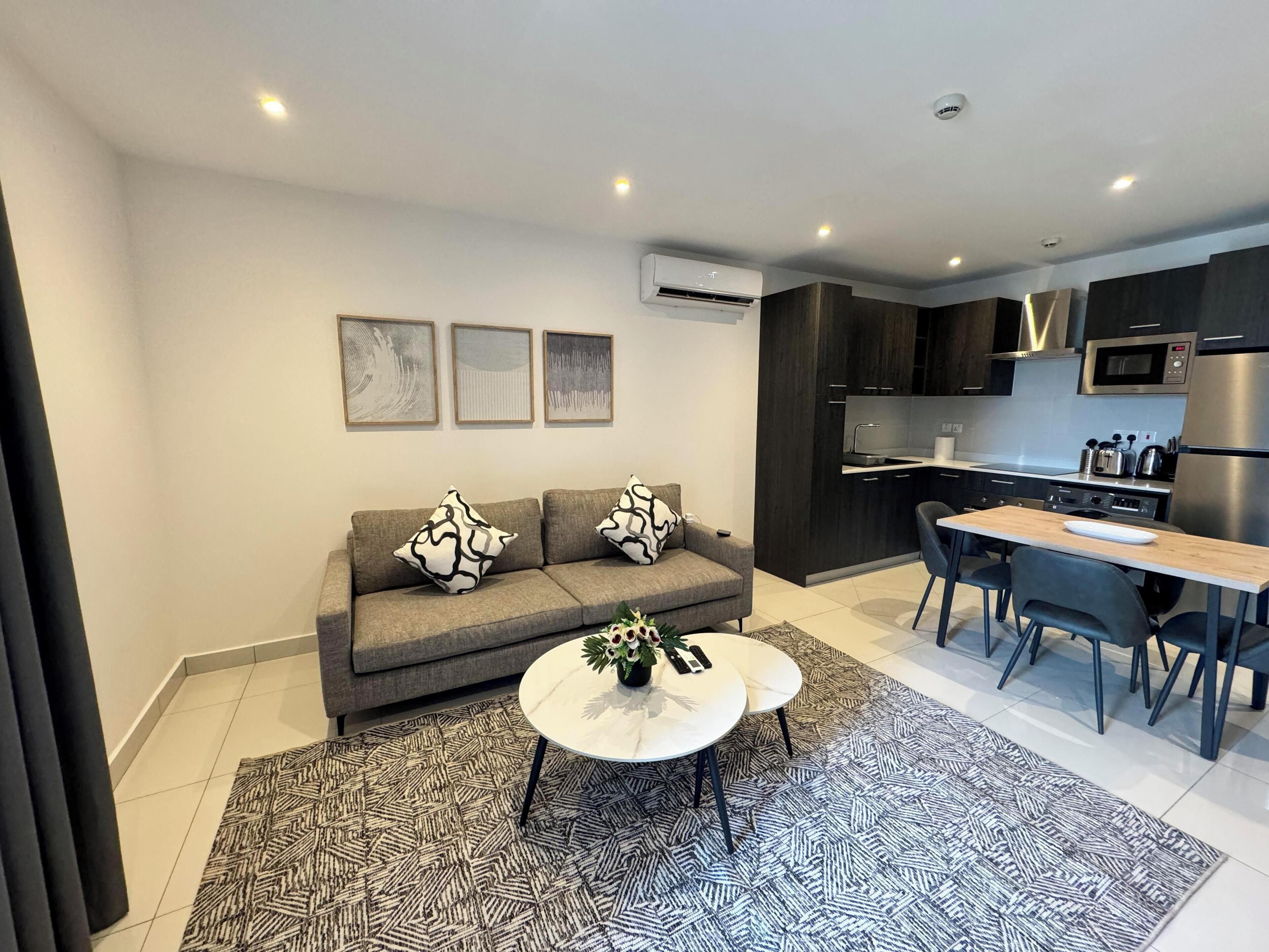 A modern living room with a grey sofa, a coffee table, and a dining area with a table and chairs. There is a kitchen with dark cabinets and stainless steel appliances.
