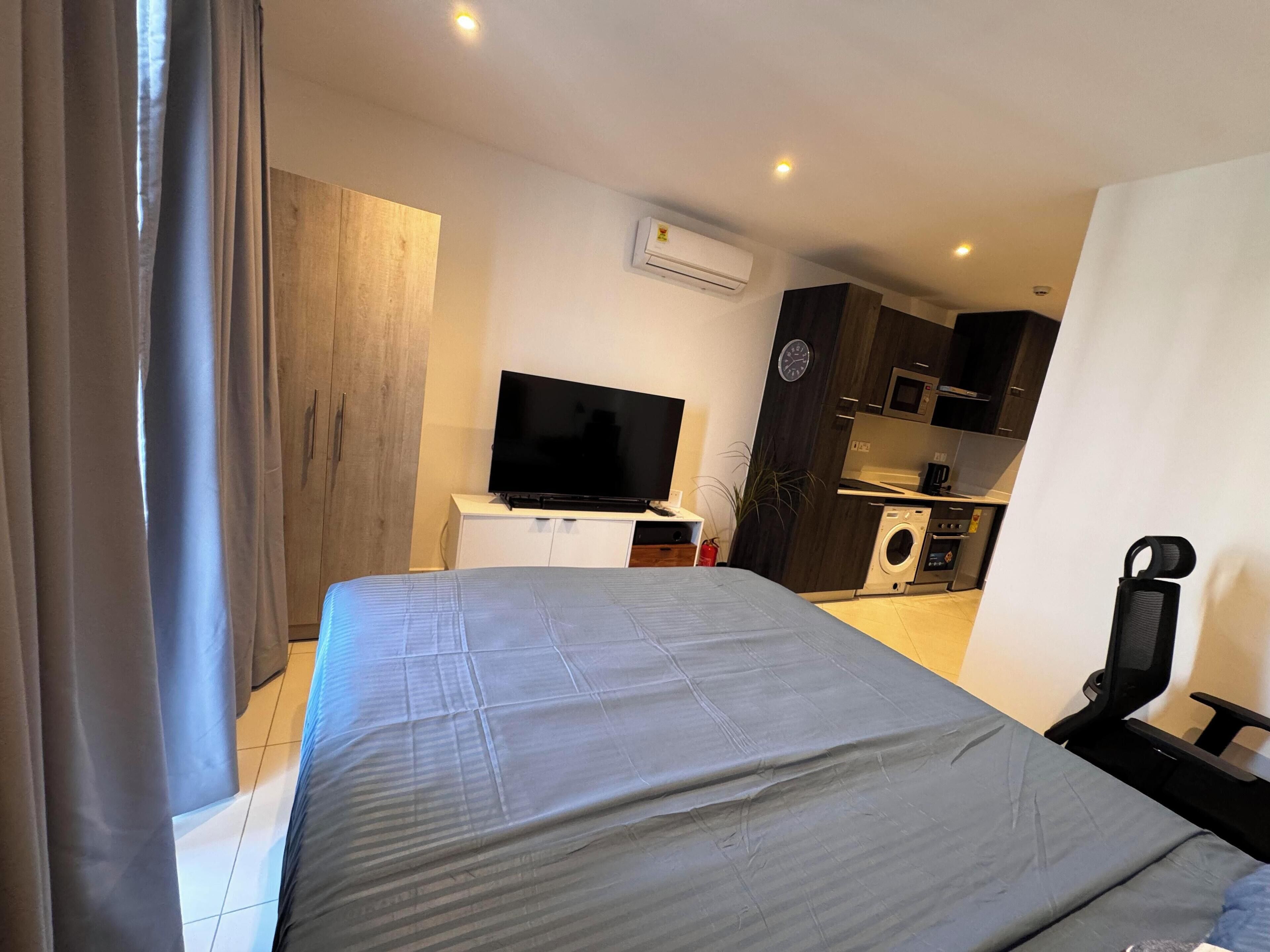A modern hotel room with a large bed, a flat-screen TV, a built-in washing machine, and a compact kitchenette.